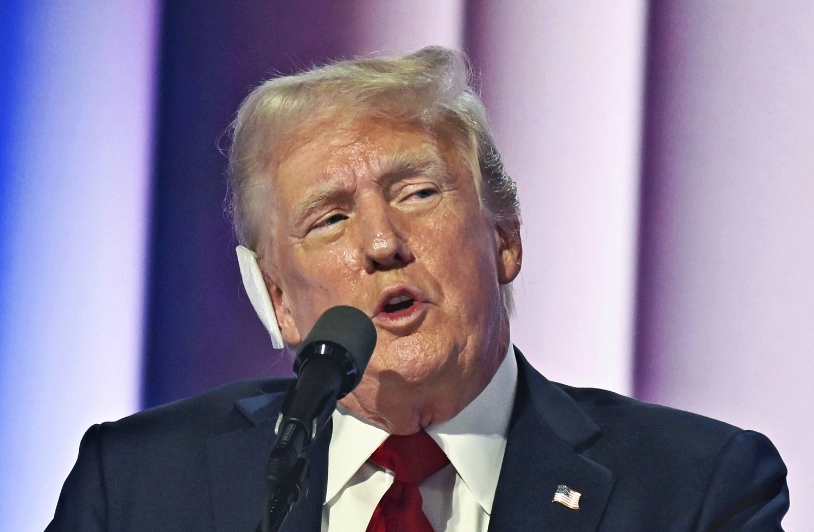 Close-up of Donald Trump speaking into a microphone during a rally, with a serious expression and a backdrop of blurred lights.