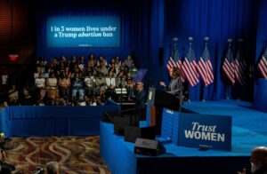 Vice President Kamala Harris speaks at a campaign event with a banner behind her reading “1 in 3 women lives under a Trump abortion ban,” emphasizing reproductive rights as a key issue in the upcoming election.