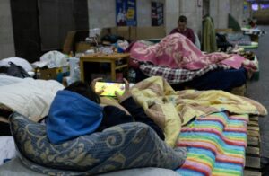 A person playing on a phone while lying on a makeshift bed in a crowded shelter, highlighting the challenges faced by evacuees during disasters like Hurricane Milton.