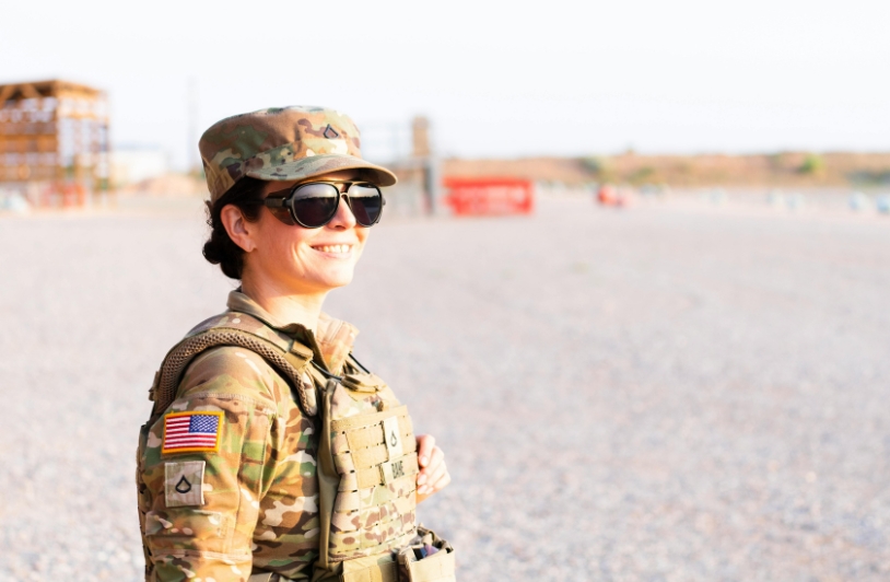 A female U.S. Army soldier wearing combat gear and sunglasses stands outdoors, smiling confidently with a blurred training field in the background.