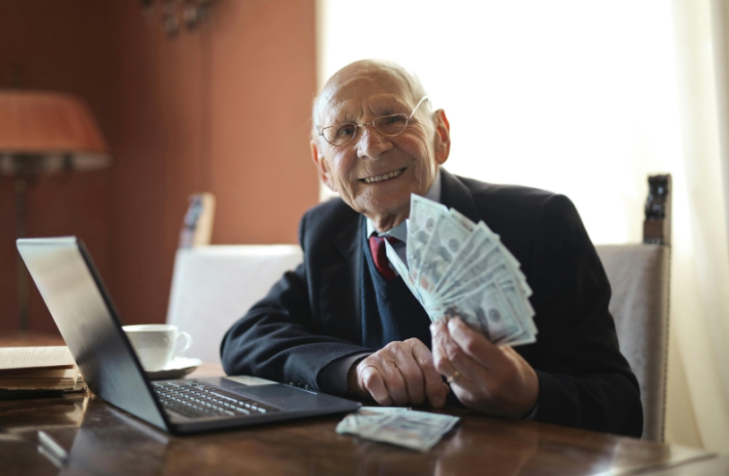 Smiling elderly man holding cash with a laptop on the table