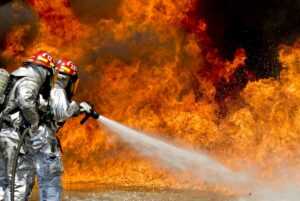 Two firefighters in reflective suits and helmets spray water onto massive flames.