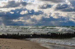 A stormy view of the Lake Superior shoreline in Upper Michigan, with waves crashing against the sandy coast and remnants of an old wooden pier beneath a dramatic cloudy sky.